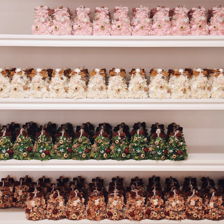 Rows of handcrafted everlasting Christmas trees made with preserved hydrangeas displayed on factory shelves, including pink and green editions before being placed in glass domes
