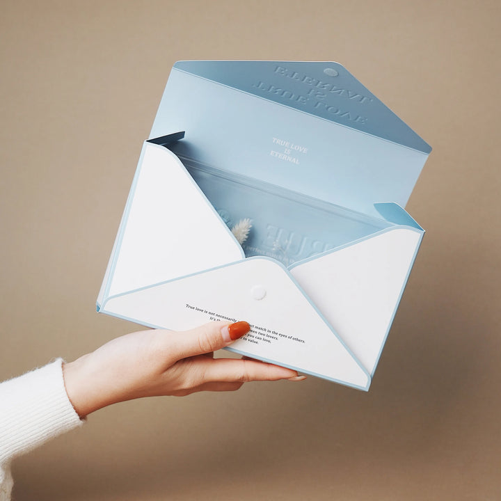 A hand holding the Everlasting Flora Envelope with blue preserved flowers and blue envelope-style box