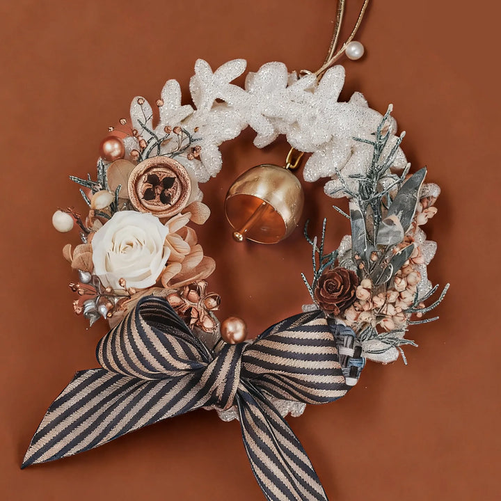 Everlasting Christmas wreath with preserved roses, hydrangeas, and festive greenery, decorated with a striped ribbon bow and golden bell on a warm brown background